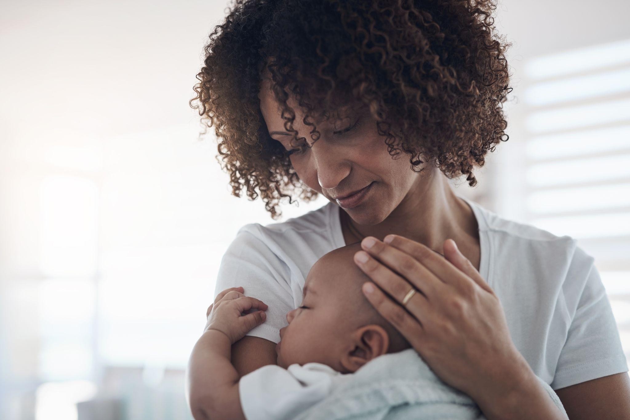 A mother with curly hair gently cradling her sleeping newborn baby against her chest.